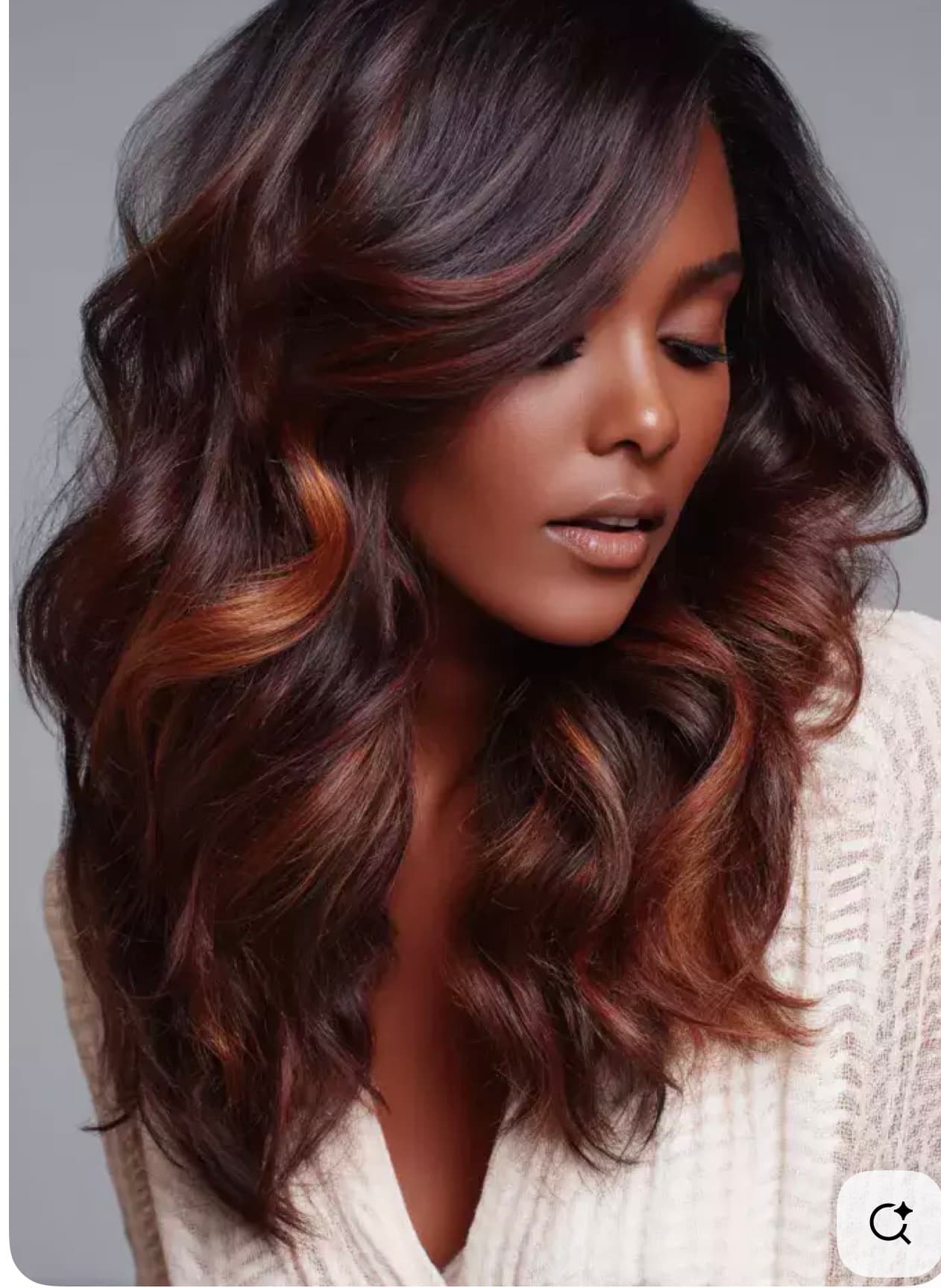 Woman with long, voluminous, wavy dark brown hair and auburn highlights looking down thoughtfully.